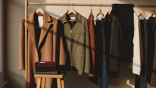 A rail of secondhand fall clothing - camel wool overcoat, olive field jacket, rust knit sweater, tweed blazer and dark denim - with folded wool scarves on a stool.