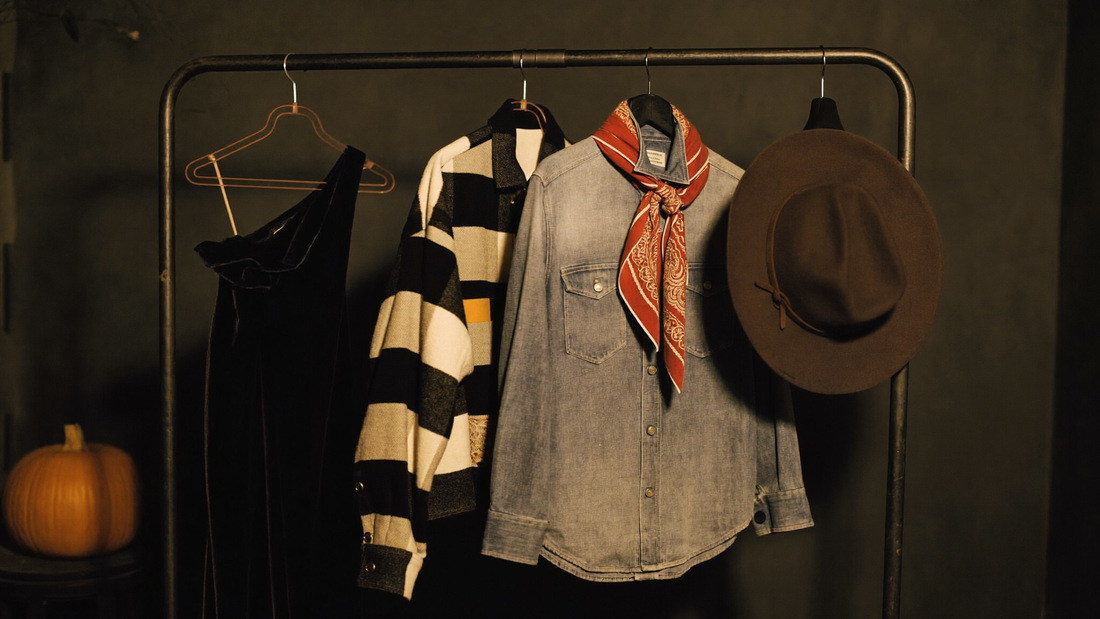 A secondhand clothing rail in warm lamplight with a black velvet dress, striped jacket, plaid flannel shirt and a wide-brim felt hat, styled for thrifted Halloween costumes.