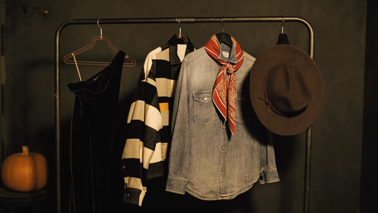 A secondhand clothing rail in warm lamplight with a black velvet dress, striped jacket, plaid flannel shirt and a wide-brim felt hat, styled for thrifted Halloween costumes.