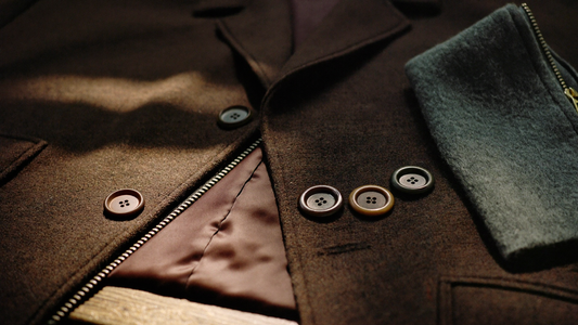 A brown tweed jacket turned inside out on a wooden table showing bound seams and silk lining, beside horn buttons, a brass zipper and a folded wool swatch.