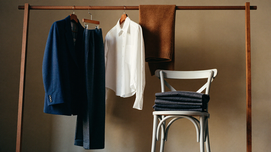 A secondhand business casual wardrobe on a wooden rail in morning light: navy wool blazer, charcoal trousers, a white cotton shirt and a camel merino knit.