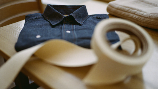 A vintage indigo denim shirt folded flat on a wooden table in soft daylight, with a plain cloth tailor's tape blurred in the foreground and a folded knit sweater beside it.