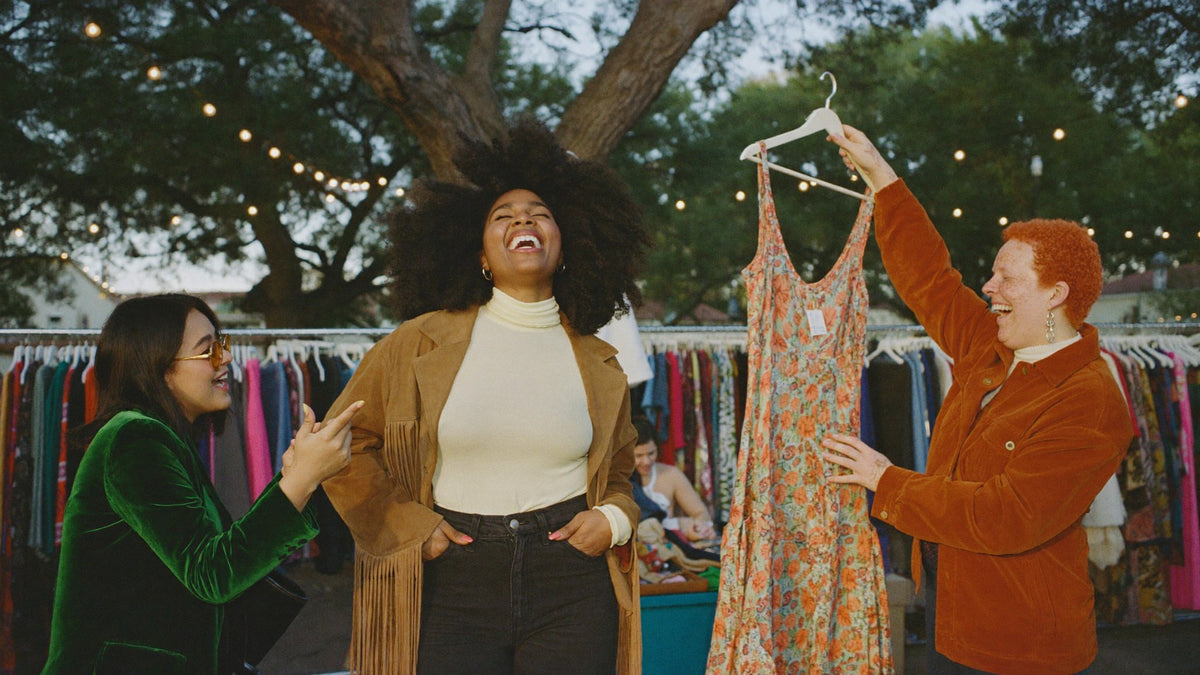 Three friends laughing while shopping racks of vintage clothing at an outdoor market under string lights